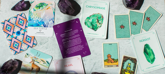 A colorful spread of oracle cards are laid across a bright background under sunlight. The cards feature crystals and their metaphysical properties, animals and their spiritual meanings, and Lenormand cards with symbolic meanings.