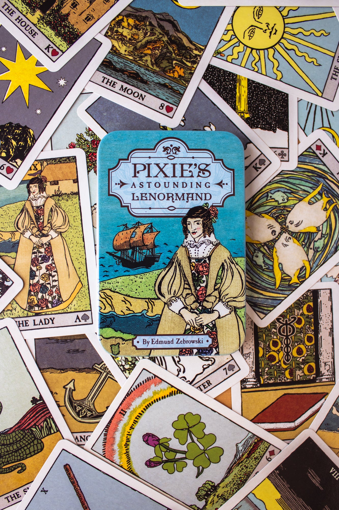 Pixie's Astounding Lenormand cards. This deck features artwork reminiscent of traditional Tarot archetypes, but in a simpler fashion. Cards are laid out in a colorful spread with the tin box sitting on top.